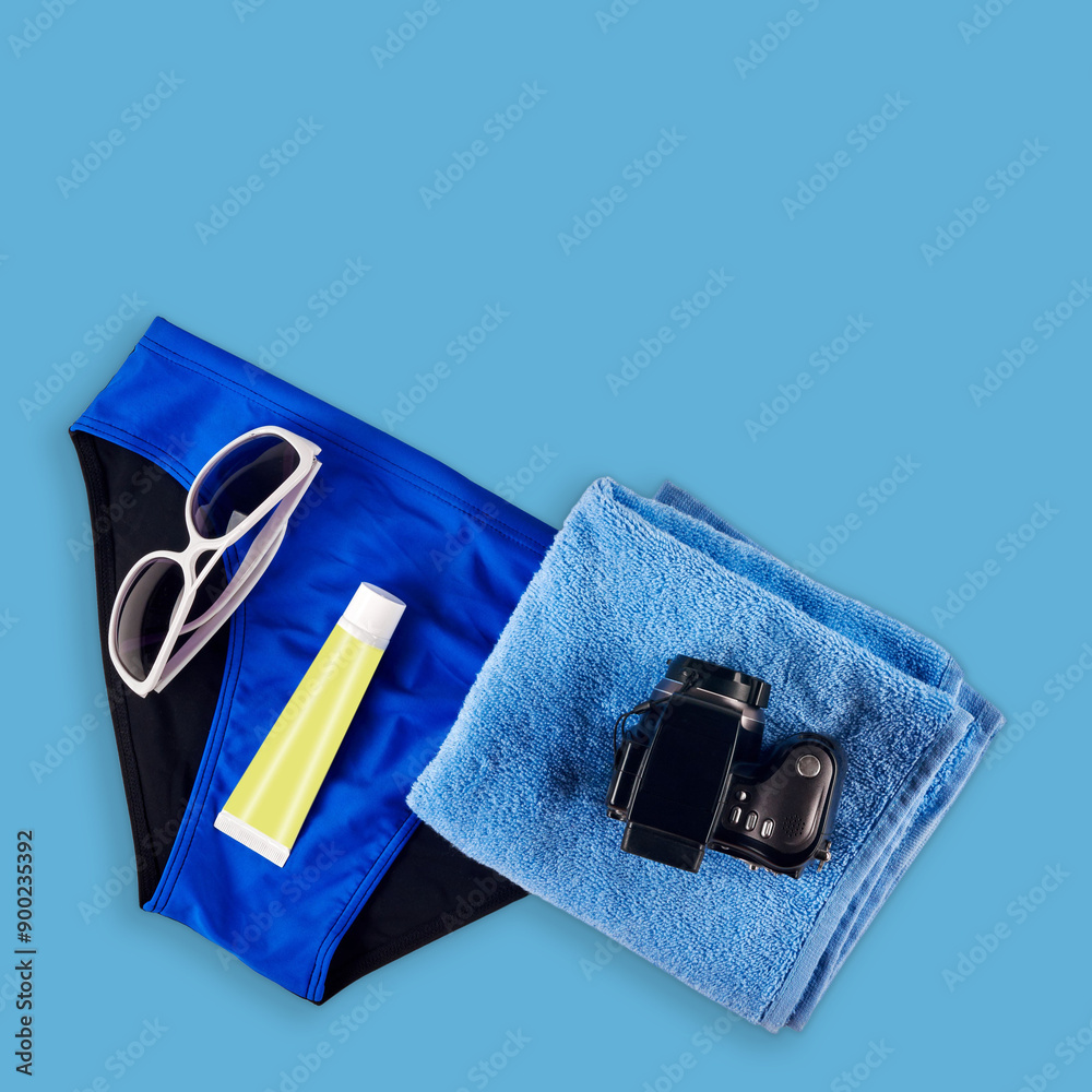 Stylish glasses, male swimsuit, modern photo camera, towel and gel on blue background. Men's beach set featuring camera, towel, sunglasses, and sunscreen, isolated Stylish glasses, male swimsuit, modern photo camera, towel and gel on blue background. Men's beach set featuring camera, towel, sunglasses, and sunscreen, isolated