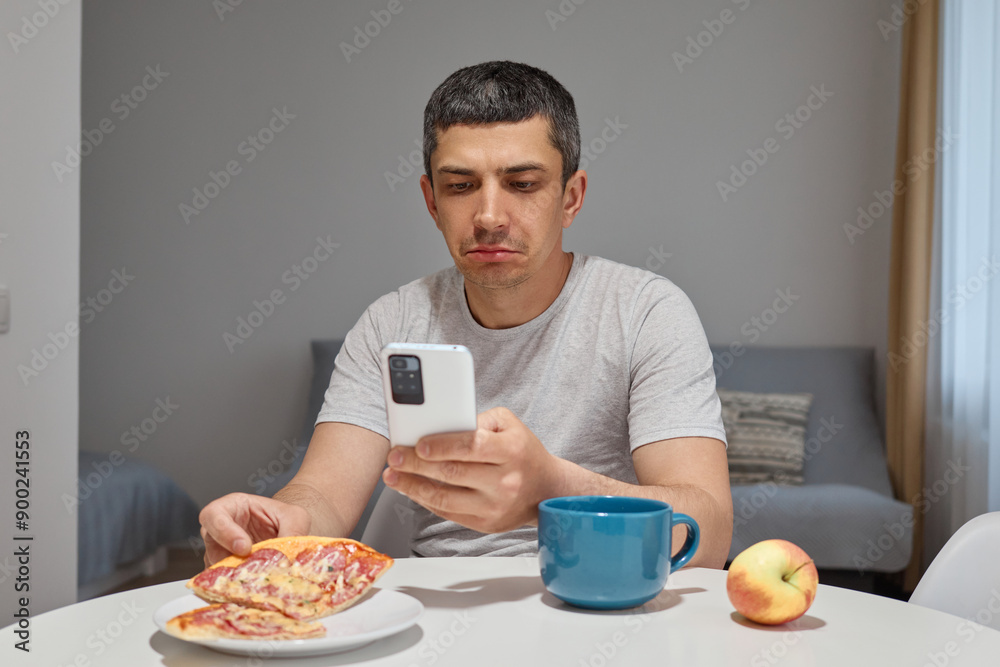 Sad upset displeased man sitting at table eating pizza and using his smartphone in home interior reading message in social network with bed news having junk snack Sad upset displeased man sitting at table eating pizza and using his smartphone in home interior reading message in social network with bed news having junk snack