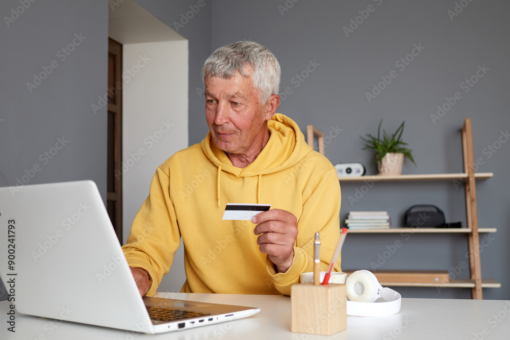 Senior Caucasian man wearing yellow hoodie sitting at home with pc computer holding credit card using e-banking services engaged in online shopping purchasing items on the internet
