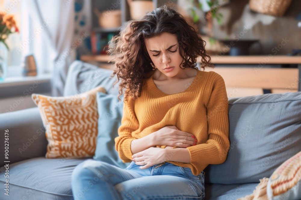 Woman in Orange Sweater Sitting on Couch with Abdominal Pain Woman in Orange Sweater Sitting on Couch with Abdominal Pain