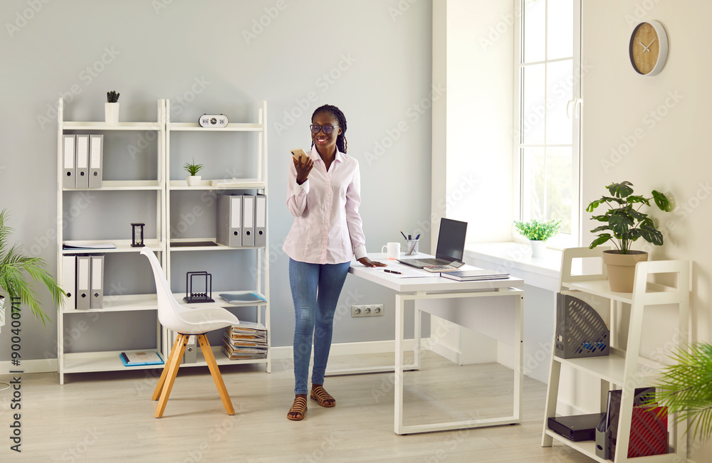 Portrait of smiling young African American office worker who using mobile phone. Dark-skinned woman is using mobile phone for messaging, using loudspeaker or browsing websites at her workplace. Portrait of smiling young African American office worker who using mobile phone. Dark-skinned woman is using mobile phone for messaging, using loudspeaker or browsing websites at her workplace.