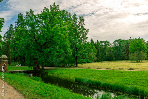 A serene path winds towards a small brick building in a lush forest, flanked by meadows and a stream. The sky is a tranquil blue with fluffy clouds, perfect for a summer day