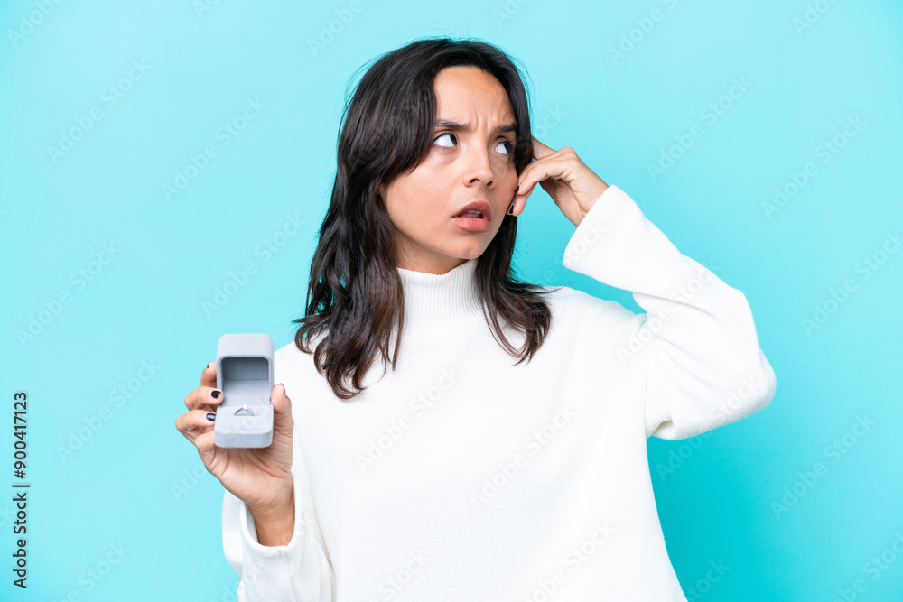 Young hispanic woman holding a engagement ring isolated on blue background having doubts and thinking