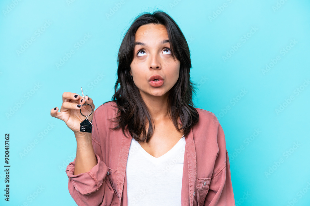 Young hispanic woman holding home keys isolated on blue background looking up and with surprised expression Young hispanic woman holding home keys isolated on blue background looking up and with surprised expression