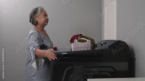 Footage Handheld medium shot, Asian mature woman holding basket with many clothes at laundry room, put dirty clothes into automatic washing machine and press to setting for start working