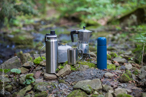 Coffee pot and metal cup on big stone against backdrop of mountain river. Coffee pot on stone plate over blurred nature background.