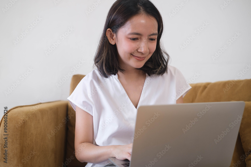 Young woman working on a laptop while sitting on a couch, focused and smiling, representing remote work or studying from home. Young woman working on a laptop while sitting on a couch, focused and smiling, representing remote work or studying from home.