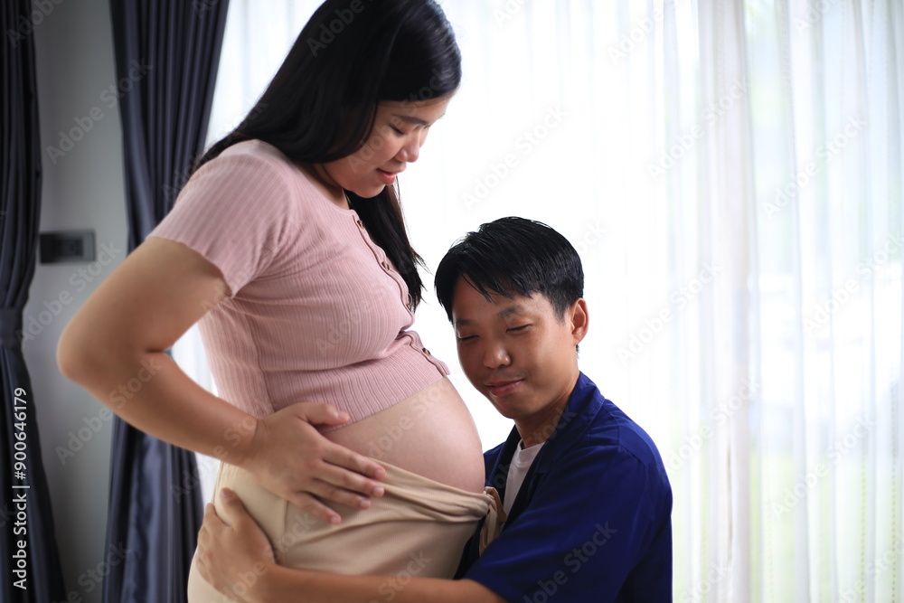 © kiattisak - Happy man with pregnant wife rubbing his belly while relaxing and spending time in modern apartment. Pregnant woman and husband spending time together using smartphone
