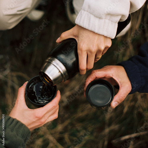 An intimate film photograph capturing hands sharing a warm drink from a thermos in a cozy outdoor setting. The image evokes feelings of friendship, warmth, connection in a serene natural environme