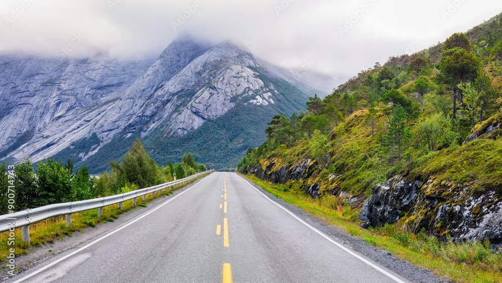 Naklejka premium Scenic mountain road lined with lush greenery and Stetind dramatic peak shrouded in mist. Ideal for nature, road trip and adventure articles and blogs . North Norway