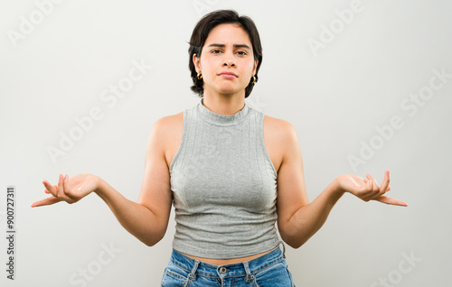 Confused young latin woman in casual attire posing with hands raised and a frown in a studio setting