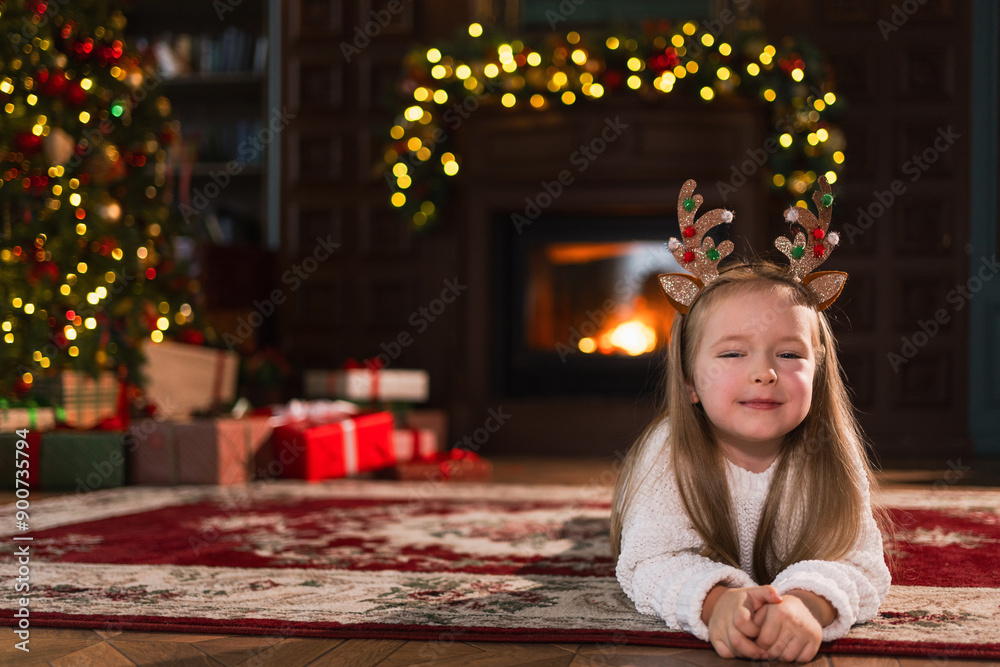 Merry Christmas. Little girl smiling near Christmas tree in classical dark interior. Young happy kid wearing deer horns in living room with fireplace Christmas tree gift boxes. Christmas eve at home Merry Christmas. Little girl smiling near Christmas tree in classical dark interior. Young happy kid wearing deer horns in living room with fireplace Christmas tree gift boxes. Christmas eve at home