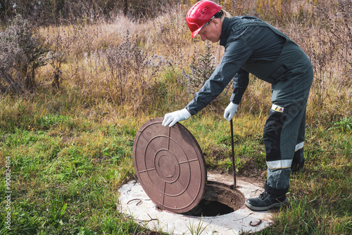 A worker in a helmet lifts a manhole cover to service a well. Plumbing and sewerage