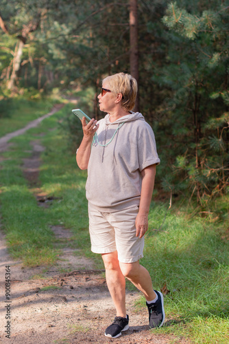 woman in the forest in beige knitted suit takes her daily steps, breathes the air of a pine forest, records an audio message on a smartphone, blogs on social networks, 5G internet photo