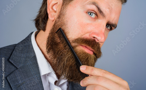 Barbershop. Portrait of brutal man with beard and mustache with barber comb. Salon for men. Stylish bearded man in suit combing beard with hair comb. Professional beard care. Barber shop advertising. photo