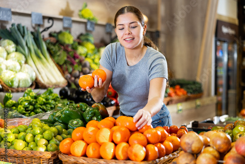 Tableau sur toile Young woman chooses tangerines for purchase in greengrocer shop