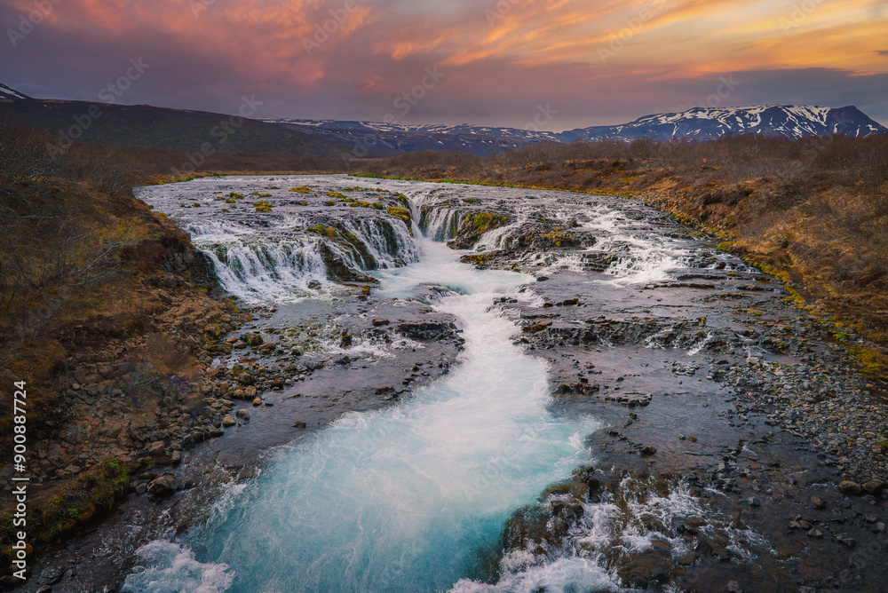 Stunning Bruarfoss waterfall in Iceland with vibrant blue waters cascading over rock formations, surrounded by rugged terrain and snow-capped mountains at sunrise or sunset. Stunning Bruarfoss waterfall in Iceland with vibrant blue waters cascading over rock formations, surrounded by rugged terrain and snow-capped mountains at sunrise or sunset.