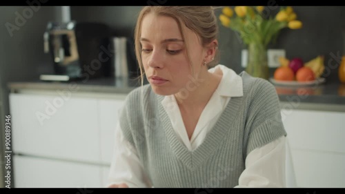 Young woman in casual white blouse and grey vest typing on laptop in a bright kitchen with modern appliances and fresh fruits, emphasizing focus and productivity in a comfortable home environment photo