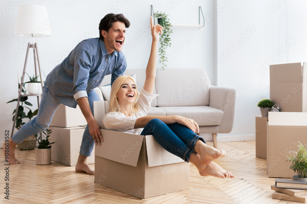 A woman sits in a cardboard box while a man pushes her through a new house, both laughing. A woman sits in a cardboard box while a man pushes her through a new house, both laughing.