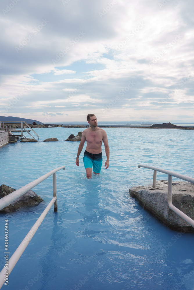 A man in blue swim trunks navigates the tranquil milky blue waters of the Blue Lagoon in Iceland, with distant mountains and volcanic rocks in the background. A man in blue swim trunks navigates the tranquil milky blue waters of the Blue Lagoon in Iceland, with distant mountains and volcanic rocks in the background.