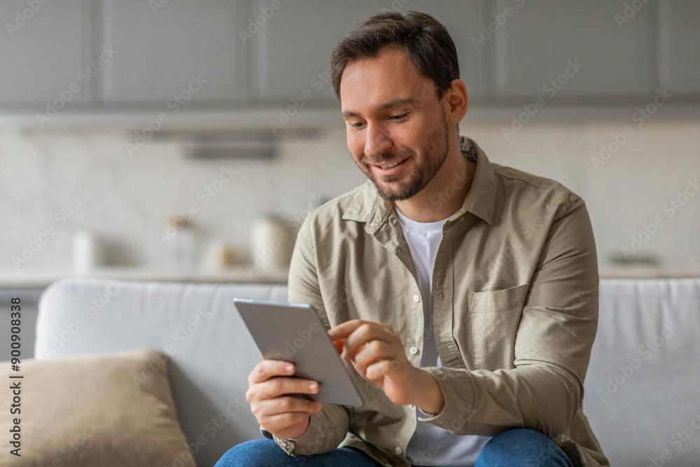 A man sits on a couch in a living room, smiling as he uses a tablet computer. He is wearing a beige button-down shirt and blue jeans.