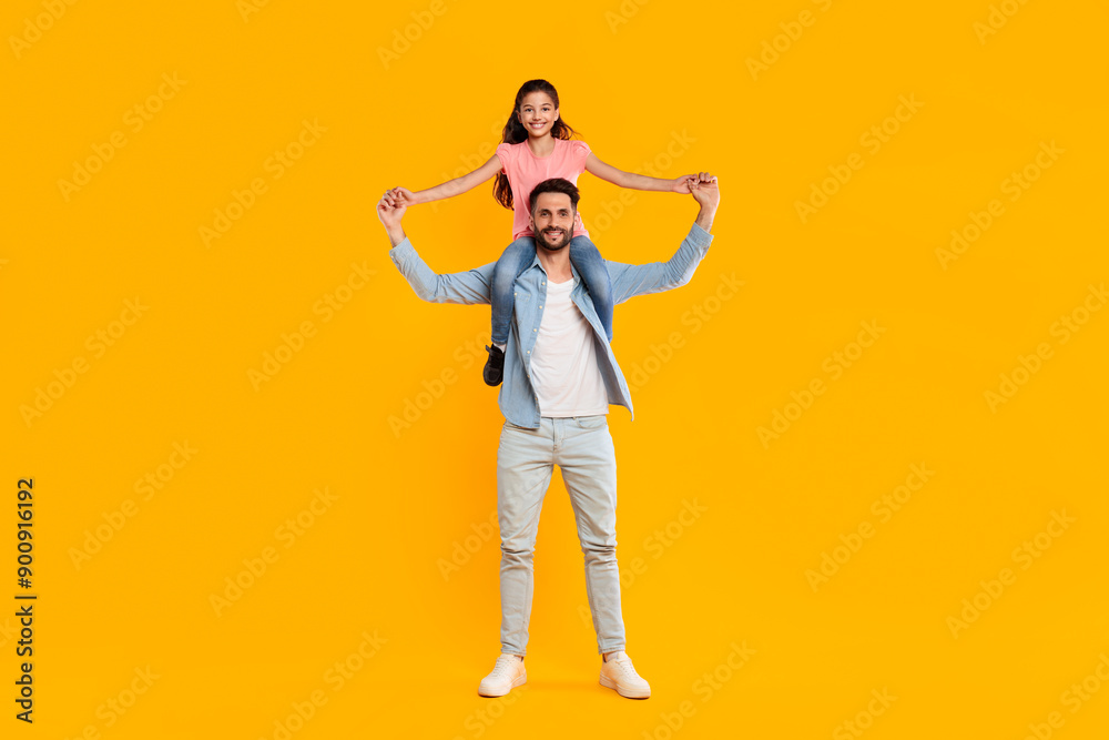 A father and his daughter are laughing together as she sits on his shoulders and holds his hands while they stand in front of a vibrant yellow background. They appear to be enjoying a playful moment. A father and his daughter are laughing together as she sits on his shoulders and holds his hands while they stand in front of a vibrant yellow background. They appear to be enjoying a playful moment.