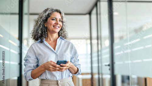 Happy middle aged business woman holding mobile cell phone using cellphone in office. Smiling mature old professional lady business investor owner entrepreneur using smartphone standing looking away.