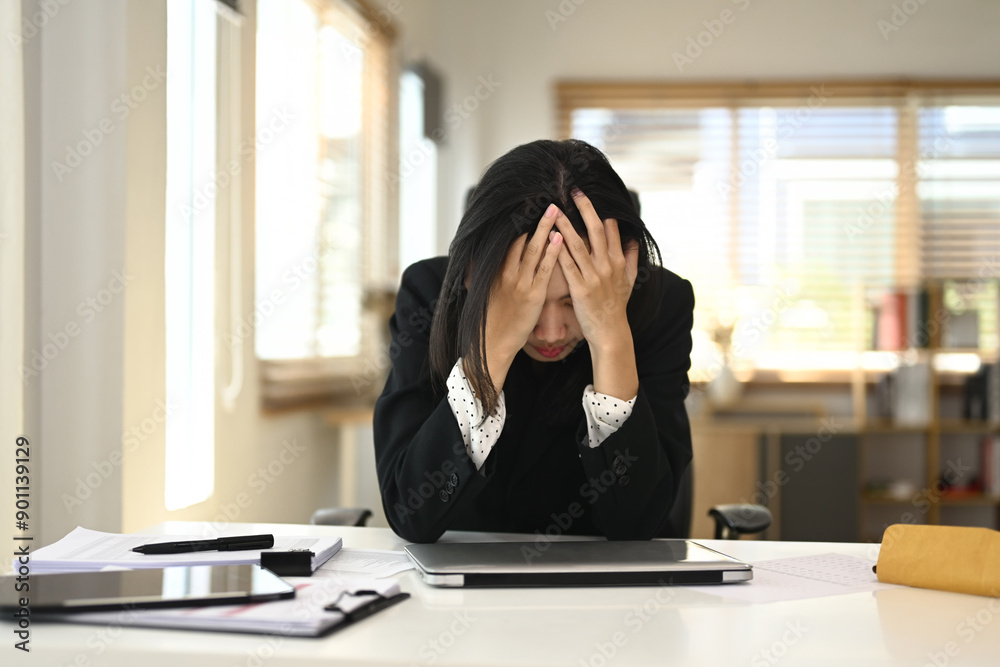 Young businesswoman sitting at office desk with head in her hands, appearing stressed or overwhelmed