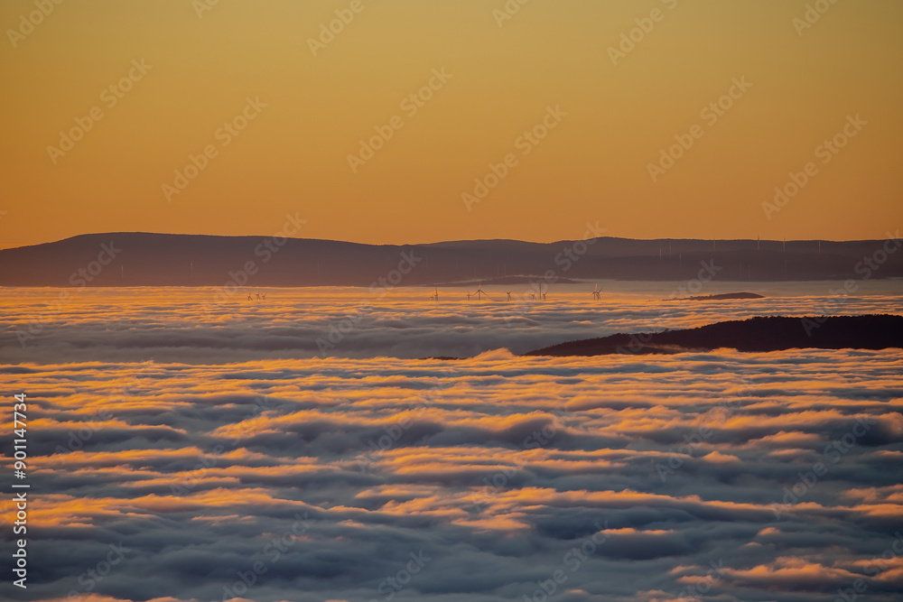 Obraz premium Cloud cover at dawn in golden rays of sun, aerial view, wind turbines in the distance above the clouds
