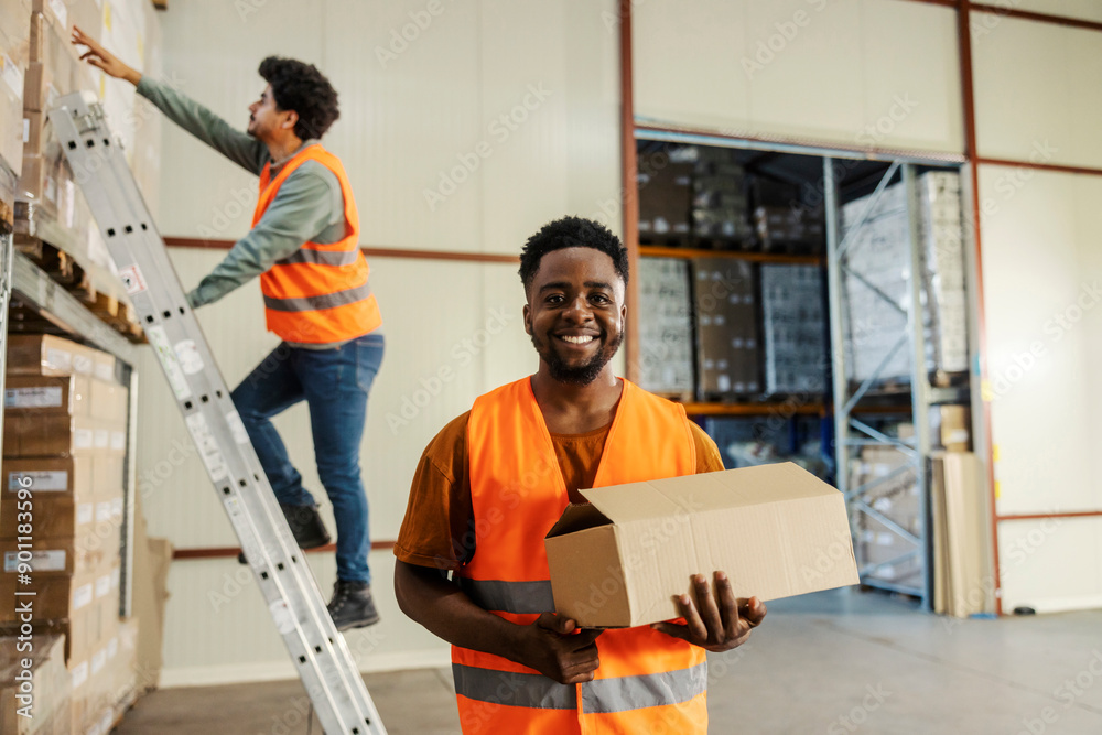 Obraz premium Portrait of diverse storage worker with box in hands smiling at camera while his coworker on ladder reaching for more boxes.