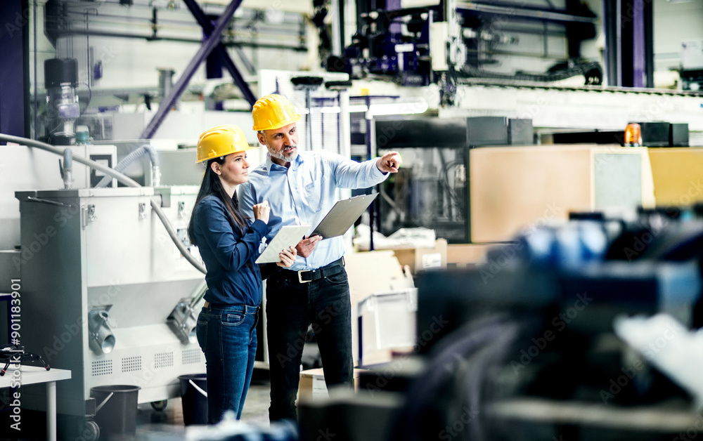 Two project managers standing in modern industrial factory, looking document in clipboard. Manufacturing facility with robotics, robotic arms and automation Two project managers standing in modern industrial factory, looking document in clipboard. Manufacturing facility with robotics, robotic arms and automation