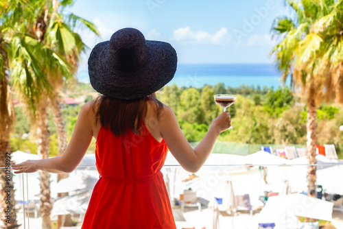 Fototapeta Naklejka Na Ścianę i Meble -  Back view of Woman in a red dress and beach hat holding cold coffee drink frappe enjoys the summer sea view, overlooking the Mediterranean Sea in Italy, Sicily. No visible face. Vacation. 