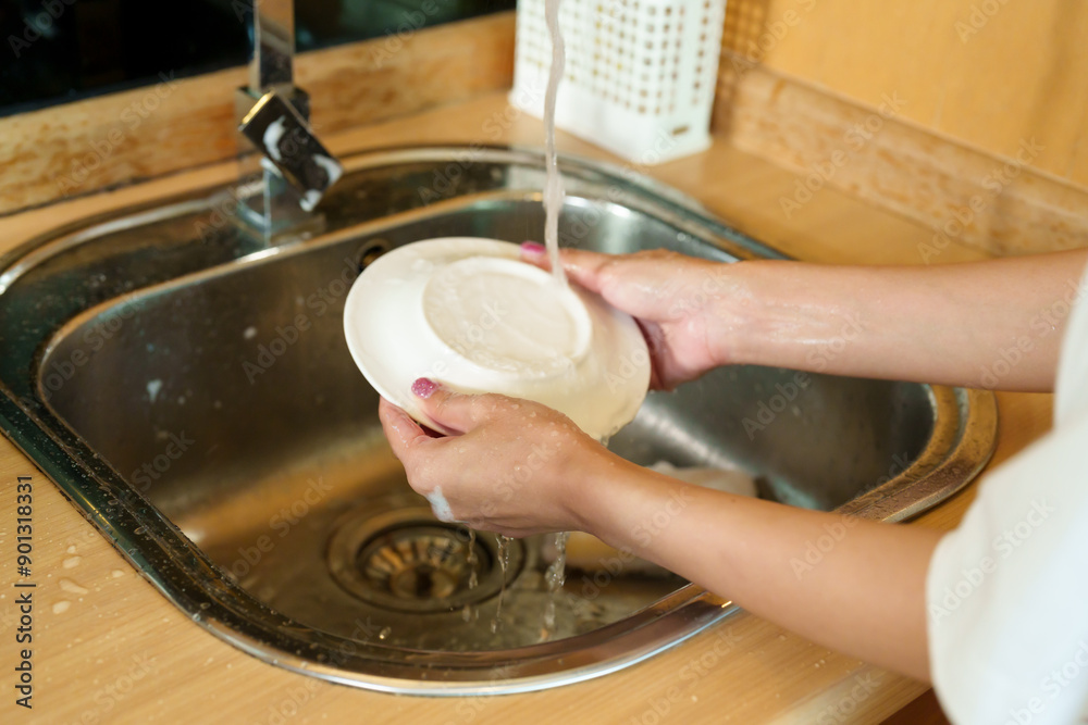 Asian woman washing dishes in the kitchen sink. scrubbing a plate with soapy sponge. Modern kitchen with wooden cabinets and stainless steel sink. Engaged in daily household chores, Asian woman washing dishes in the kitchen sink. scrubbing a plate with soapy sponge. Modern kitchen with wooden cabinets and stainless steel sink. Engaged in daily household chores,