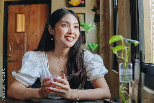 Quadro su tela Beautiful Asian woman is sitting in a cafe while holding a glass of tea, smiling to the camera, nature cafe background