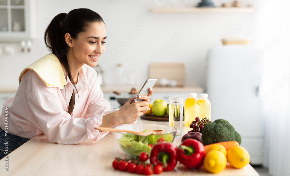 Smiling young woman using cellphone while cooking dinner in kitchen at home, cheerful casual female leaning on table, reading text messages on her cell phone and smiling, free copy space Smiling young woman using cellphone while cooking dinner in kitchen at home, cheerful casual female leaning on table, reading text messages on her cell phone and smiling, free copy space