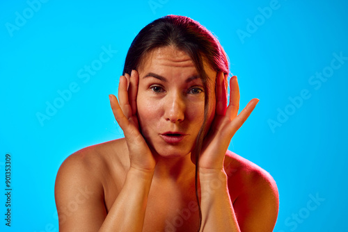 Woman with glowing skin places her hands on her cheeks, emphasizing her smooth complexion and vibrant appearance in neon light against blue background. Concept of skincare, cosmetology, dermatology. photo