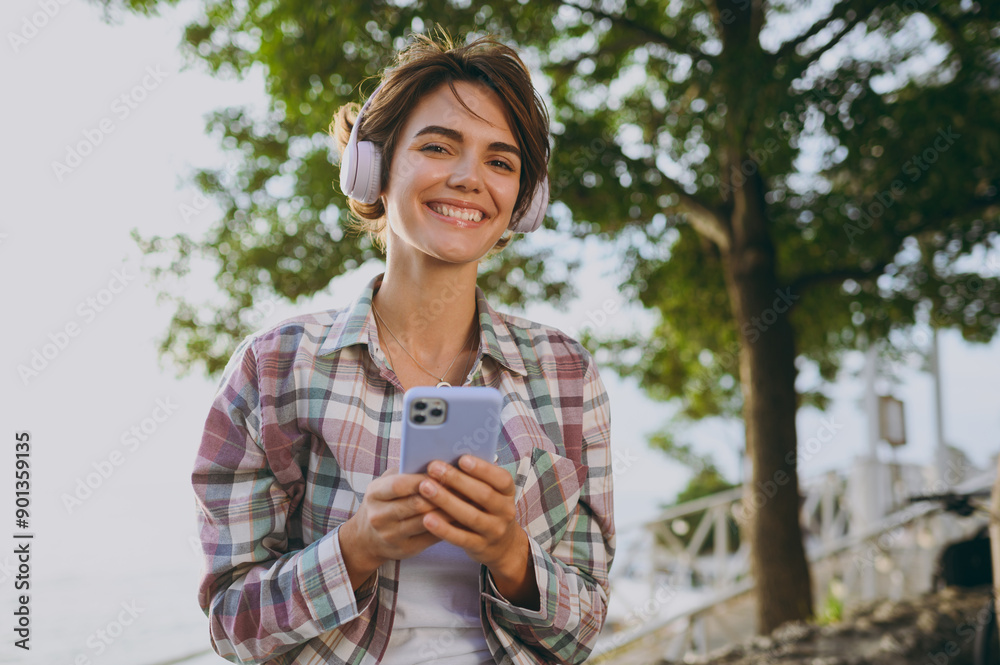 Fototapeta premium Young fun woman wear shirt casual clothes headphones hold use mobile cell phone listen music sits on bench rest in park near sea ocean beach outdoor seaside in summer day free time. Lifestyle concept.