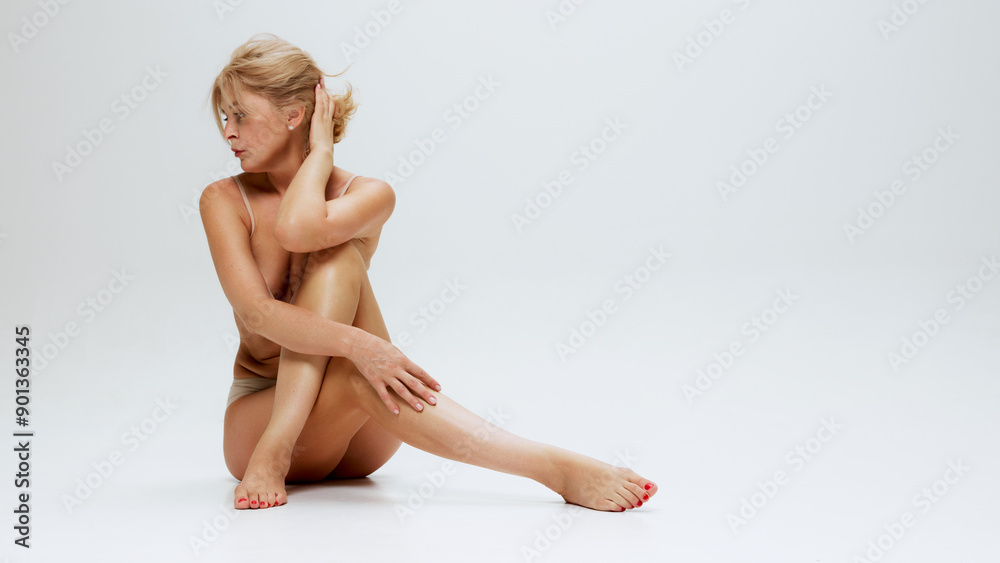 Obraz premium Seated gracefully on floor, beautiful blonde woman in lingerie, exuding serenity and confidence against white studio background. Concept of beauty, lifting, cosmetology procedures, antiaging. Ad