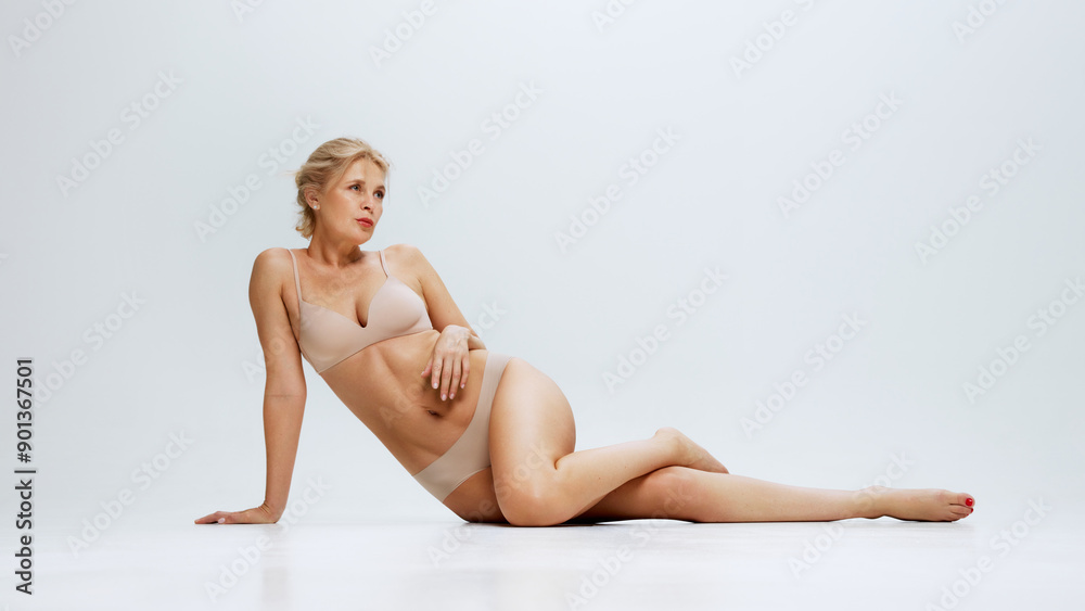 Obraz premium Elegant woman in beige lingerie lying, posing on floor against white studio background, exuding beauty an femininity. Concept of beauty, lifting, cosmetology procedures, body care, antiaging. Ad