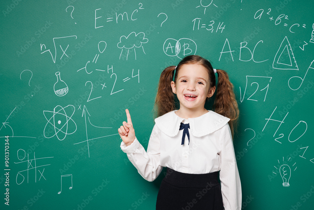 Little fun school girl 7-8 years old pony tails wear white shirt uniform point index finger indicate on area isolated on green blackboard background studio. Childhood kids education lifestyle concept. Little fun school girl 7-8 years old pony tails wear white shirt uniform point index finger indicate on area isolated on green blackboard background studio. Childhood kids education lifestyle concept.