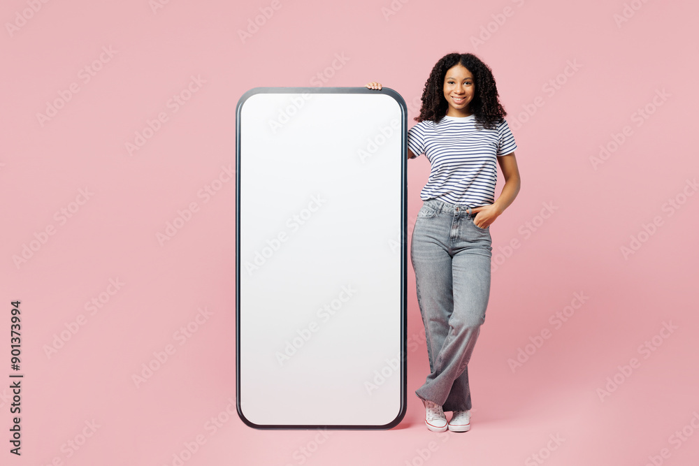 Full body little kid teen girl wearing blue t-shirt casual clothes stand near big huge blank screen mobile cell phone smartphone with area isolated on plain pastel pink background. Childhood concept. Full body little kid teen girl wearing blue t-shirt casual clothes stand near big huge blank screen mobile cell phone smartphone with area isolated on plain pastel pink background. Childhood concept.