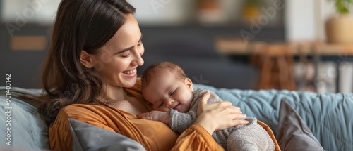Wallpaper Mural A smiling mother with long hair cradles a sleeping baby on a grey couch. The cozy background includes a bookshelf, a chair, and a plant, radiating a tranquil atmosphere. Torontodigital.ca
