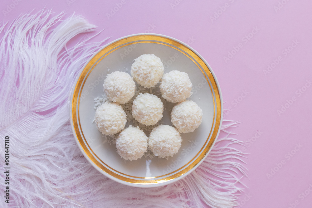 Top view of White Chocolate Candy With Coconut Topping On White Feather and pink background, sweet balls on plate flat lay with copy space. Romantic gift for celebration or anniversary