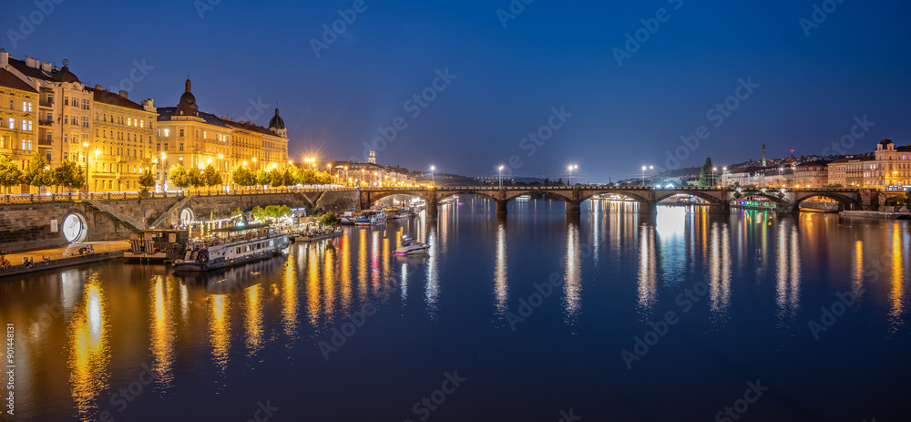 Obraz premium A view of the Rasin Embankment and Palacky Bridge in Prague, Czechia at dusk. The bridge is lit up, and the city lights reflect in the water. There are boats on the river, and the sky is a deep blue.