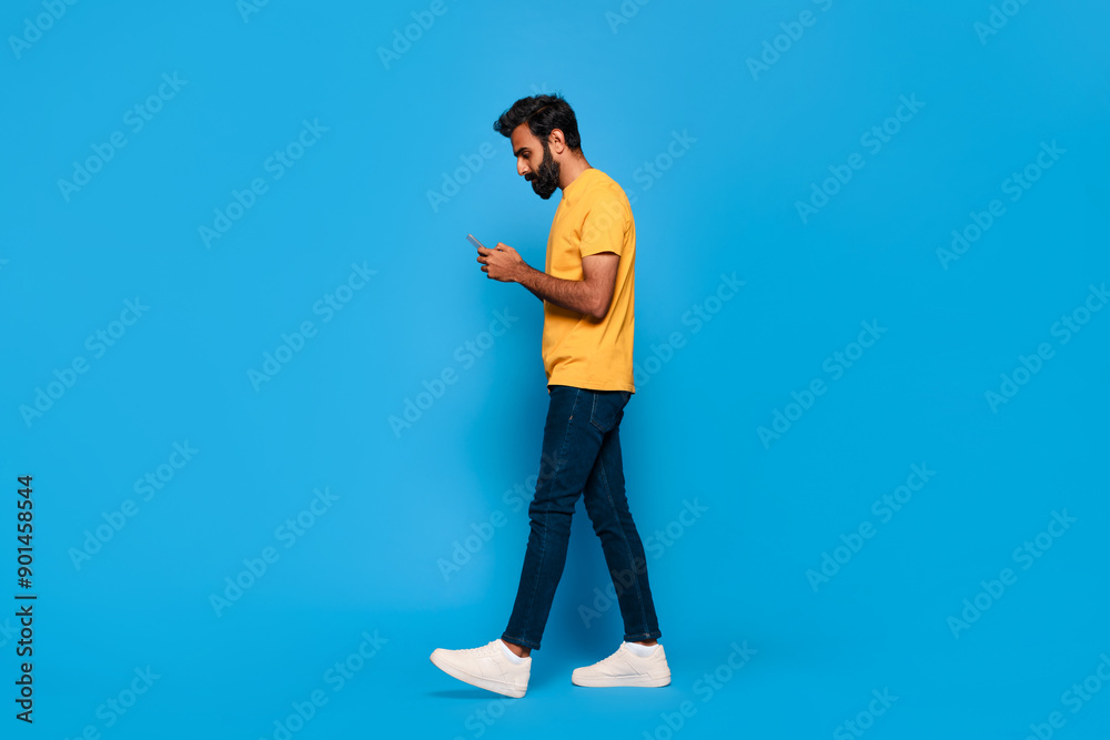 Indian man walks while looking at his phone, smiling slightly. He is wearing a yellow t-shirt, blue jeans, and white sneakers. The background is a solid blue. Indian man walks while looking at his phone, smiling slightly. He is wearing a yellow t-shirt, blue jeans, and white sneakers. The background is a solid blue.