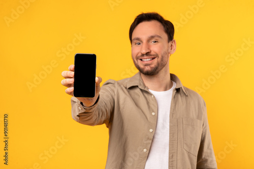 A man wearing a light brown button-down shirt smiles at the camera as he holds a smartphone with a black blank screen out in front of him. The background is a solid yellow.