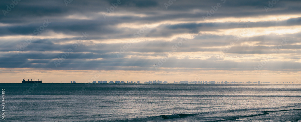 Fototapeta premium A lone ship sails on the calm ocean, with a distant city skyline visible on the horizon under a dramatic, cloudy sky.