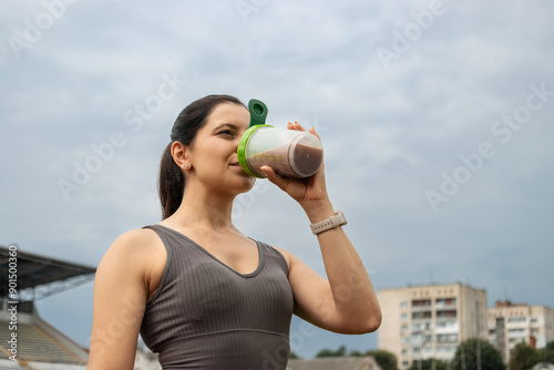 A young sporty woman drinking protein smoothie, milkshake from a shaker, resting after morning workout on the stadium, outdoors photo