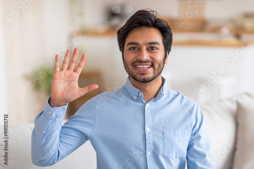 Arab man in a light blue shirt smiles and waves hello with his right hand, standing indoors in a home setting with a white couch partially visible in the background. photo