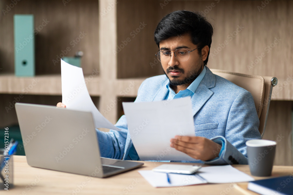 Indian businessman wearing glasses and a blue blazer sits at his desk, intently reading documents while using a laptop, desk is cluttered with papers and office supplies, indicating a busy workday. Indian businessman wearing glasses and a blue blazer sits at his desk, intently reading documents while using a laptop, desk is cluttered with papers and office supplies, indicating a busy workday.